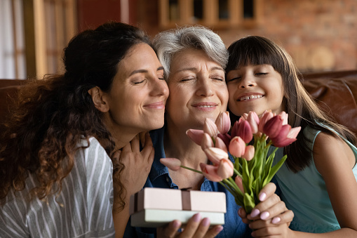 Close up happy three generations of women celebrating event, touching cheeks, cute little girl with young mother congratulating grandmother with birthday or 8 march, presenting gift and flowers