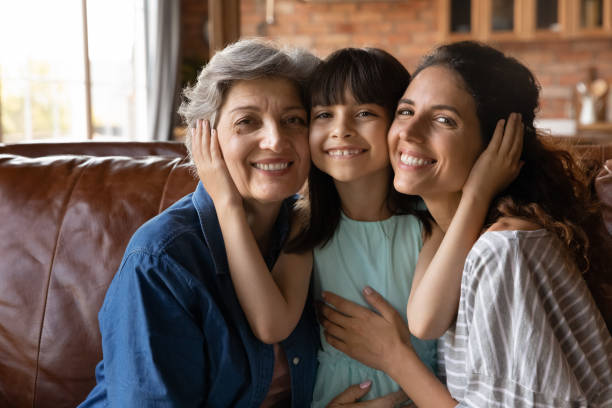 Head shot portrait happy three generations of women hugging Head shot portrait happy three generations of women hugging, touching cheeks, smiling little girl sitting on couch between young mother and mature grandmother, posing for family photo at home granddaughter photos stock pictures, royalty-free photos & images