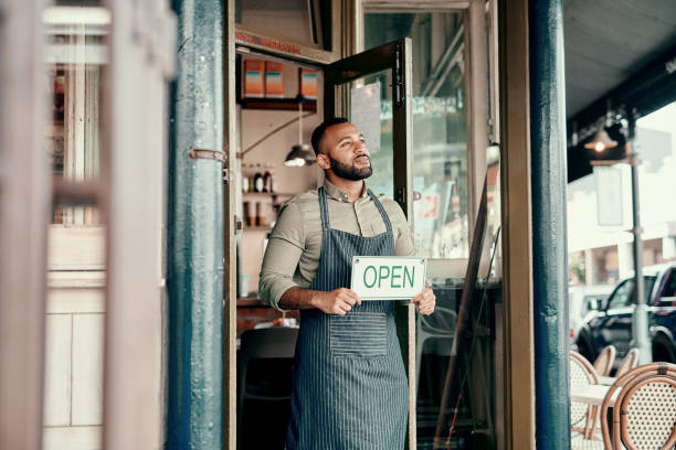 Day one of my dream come true Shot of a young man holding an "open" sign at the doorway of his cafe franchising photos stock pictures, royalty-free photos & images