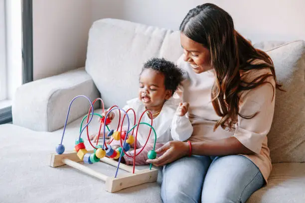 Photo of Happy smiling mixed race Indian mother and African black baby toddler playing developmental wooden bead maze toy game together at home. Authentic candid lifestyle with infant kid child.