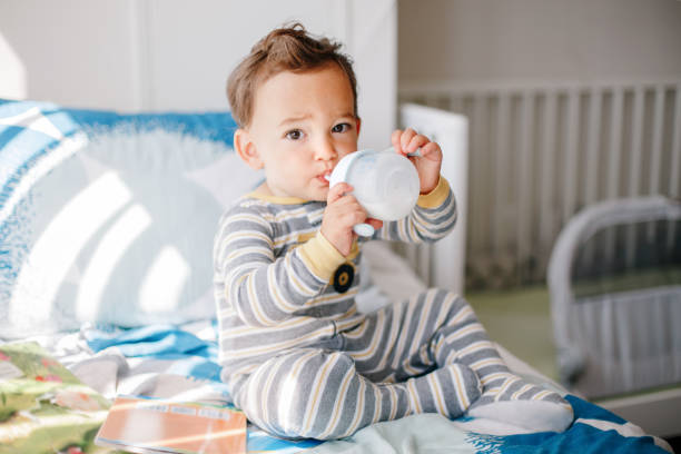 cute adorable caucasian kid boy sitting on bed drinking milk from kids bottle. healthy eating drinking for children. supplementary food for growing babies. candid real authentic moment. - criança de 1 a 2 anos imagens e fotografias de stock