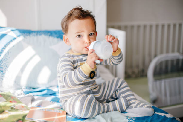 um adorável garoto caucasiano sentado na cama bebendo leite da garrafa infantil. alimentação saudável bebendo para crianças. alimentos suplementares para bebês em crescimento. momento autêntico sincero. - 18 a 23 meses - fotografias e filmes do acervo