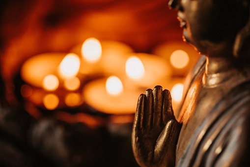 Close up of a buddha figurine seen in profile slightly in front of lit candles. Focus on the hand. For mindfulness and wisdom concepts.  Selective focus and added grain. Part of a series.