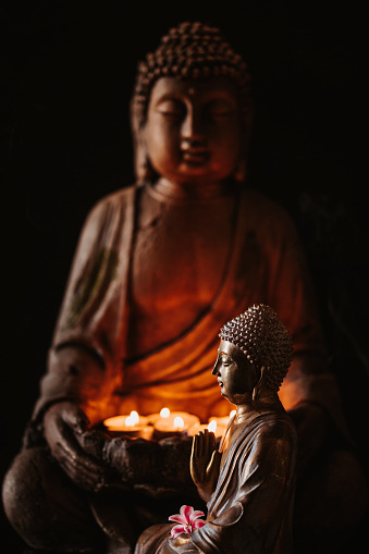 Buddha figurine seen in profile with a flower in front of another buddha holding lit candles in the hands. For mindfulness and wisdom concepts.  Selective focus and added grain. Part of a series.