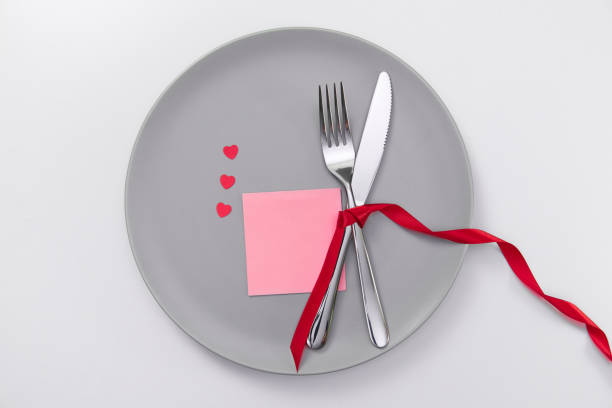 istock Valentine's day festive table setting card for text, flat lay with red heart shape on gray plate, fork, knife and red ribbons on white table. Love dating concept. Place your text and copy space 1300386264