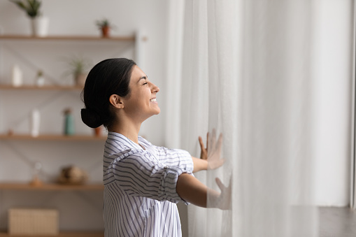 Bathing in sunlight. Excited young indian lady meet first morning at new flat house part curtains enjoy being homeowner. Happy hindu female open drapes on window breath fresh air close eyes in delight