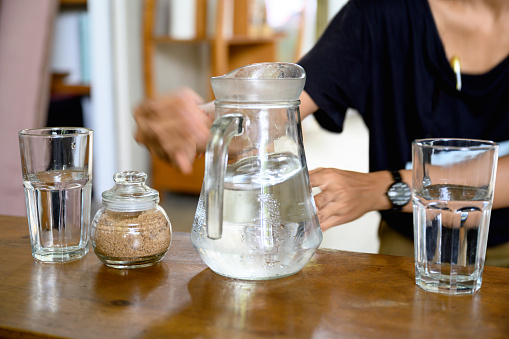 Unrecognizable woman picking up jug filled withcold water, two drinking glasses are on table, Nikon Z7