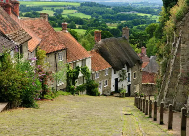 View To Old Limestone Houses On A Cobble Street At Gold Hill In Shaftesbury England With A Beautiful Rural Landscape In The Background On An Overcast Summer Day