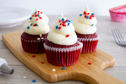 Three red velvet cupcakes with vanilla buttercream icing decorated with red, white and blue star sprinkles.
Shot on wooden chopping board in a bright kitchen.
Close up.