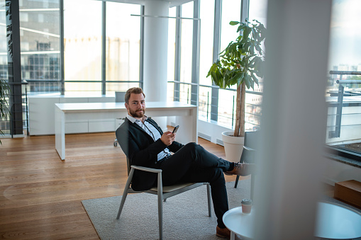 Young businessman photographed through window while sitting in office lobby, checking smart phone, and looking at camera.