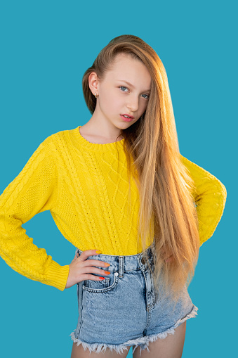 Kids fashion. Casual beauty. Modern childhood. Portrait of confident young model girl with long fair hair in bright yellow sweater denim shorts standing in akimbo position isolated on blue background.
