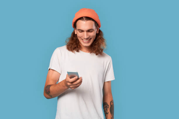Freestyle. Curly-haired guy wearing hat standing isolated on blue browsing social media on smartphone happy