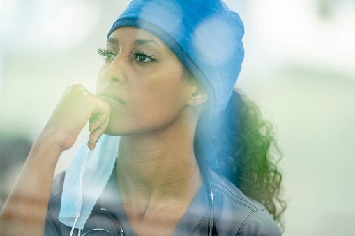 A female nurse of African descent appears to be sitting down and occupied with her thoughts. She has her hand on her chin and is looking down on the ground. Her face mask is hanging on one ear.