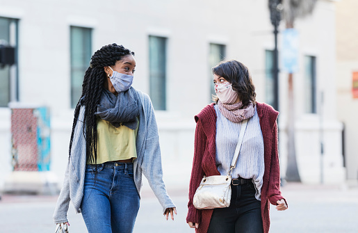Two multi-ethnic young women in their 20s walking across a city street. They are side by side, looking at each other, conversing. They are wearing protective face masks, trying to prevent the spread of coronavirus during the COVID-19 pandemic.
