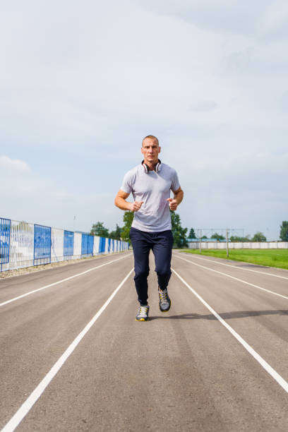 vue avant pleine longueur sur le jogging caucasien adulte d’homme sur la piste courante - athlète masculin blond dans la course d’entraînement de stade dans le printemps ensoleillé ou le jour d’été - concept sain de mode de vie de vraies personn - track and field 30s adult athlete photos et images de collection