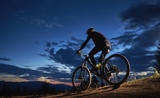 Back view of young man cycling bicycle under beautiful night sky. Male bicyclist in safety helmet riding on hillside trail under blue cloudy sky at night. Concept of sport, biking and active leisure.