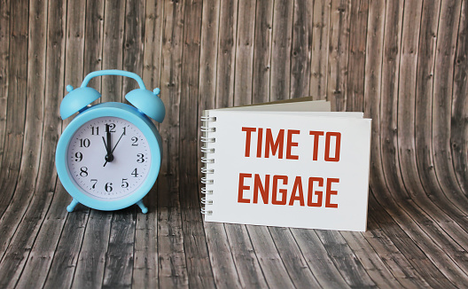 Text, the word Time To Engage is written in a notebook lying on a wooden table with an alarm clock. Business concept