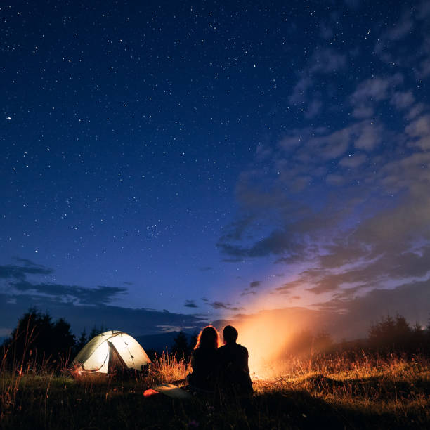 istock Young couple sitting near campfire under cloudy night sky. 1300436913