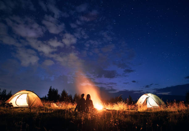 istock Young couple sitting near campfire under cloudy night sky. 1300436921