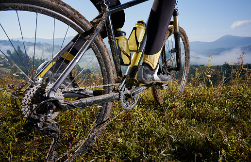 Close up, low angle view on a bike, back wheel on foreground. Man's legs riding his bicycle on grass in summer in the mountains against blue sky. Concept of extreme cycling