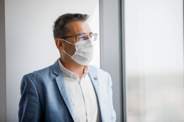 istock Businessman with protective face mask standing by the office window and looking pensive 1300444357