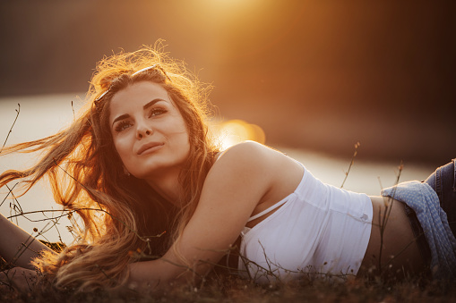 One beautiful young woman with long hair lying on her stomach on grassy  lakeshore and enjoying summer sunset over the lake
