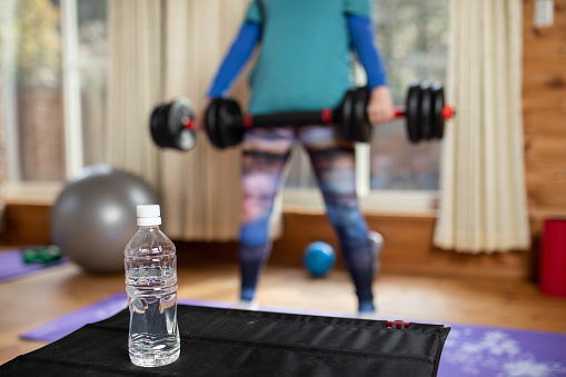 A water bottle in focus in the foreground with a woman out of focus in the background working out at home in her living room.