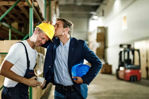 Mid adult inspector kissing manual worker's hardhat who is having a coffee break in distribution warehouse.