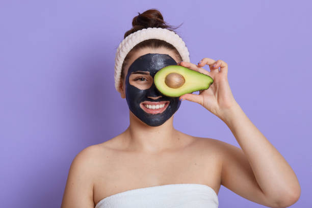 Young woman with smile holding avocado and covering eye with it, looking directly at camera, has good mood while doing beauty and cleansing procedures, girl with hair band against lilac wall. Young woman with smile holding avocado and covering eye with it, looking directly at camera, has good mood while doing beauty and cleansing procedures, girl with hair band against lilac wall. woman applying wallcovering stock pictures, royalty-free photos & images