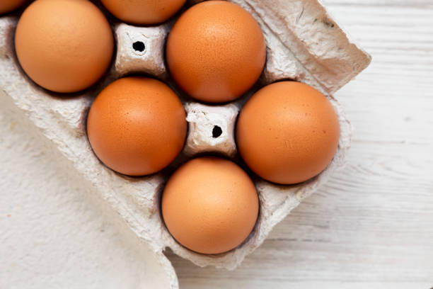 istock Raw Brown Eggs in a paper box on a white wooden background, top view. Flat lay, overhead, from above. 1300469095