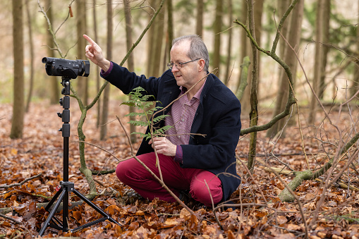 Caucasian man of 56 sitting in the forest at winter, operating a camera while vlogging about nature themes such as botany, biology and environmental conservation.