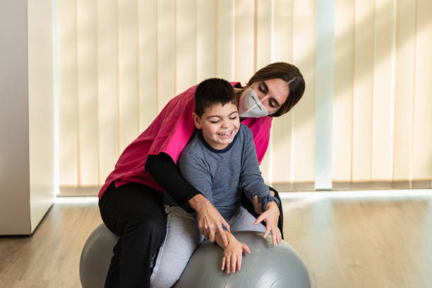 niño discapacitado y fisioterapeuta en la parte superior de un peanut gym ball haciendo ejercicios de equilibrio. protección contra máscaras pandémicas - brain gym fotografías e imágenes de stock