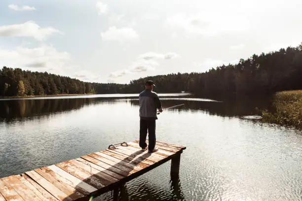 September 19, 2020 - Druskininkai, Lithuania: healthy active senior man standing alone on a jetty on a sunlit lake and fishing - holding his fishing rod, back turned, being calm, content, serene.