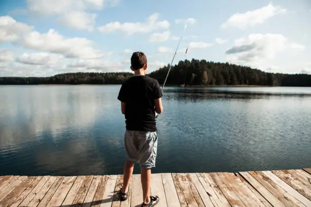 September 19, 2020 - Druskininkai, Lithuania: teenager boy holding fishing rod, standing on a jetty on a lake, catching fish, rear view, obscured face, sunny autumn day, calm waters.