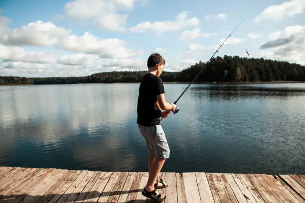 September 19, 2020 - Druskininkai, Lithuania: side view of a teen boy holding a fishing rod backlit on a lake jetty, waiting for a fish to catch the bait, being calm in a serene nature.