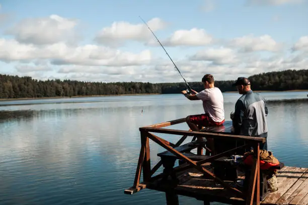 September 19,  2020 - Druskininkai, Lithuania: Grandfather and grandson idyllic togetherness and bonding - active senior man helping and teaching his teen boy fishing on a calm sunny lake sitting on a jetty, giving a hand and advice. Two generation men bonding.