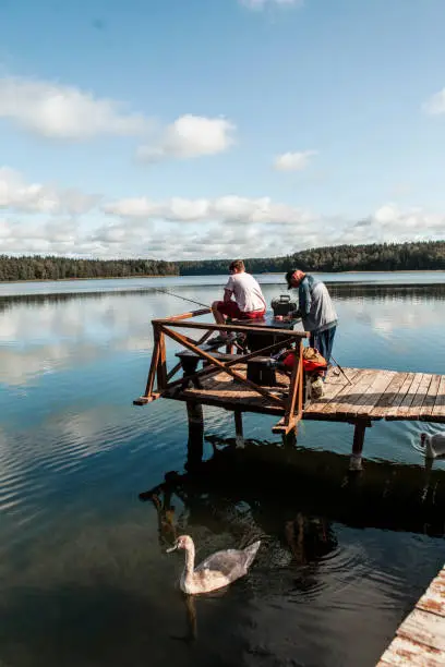September 19,  2020 - Druskininkai, Lithuania: grandpa helping and giving advice his teen grandson on fishing. They sit on a wooden jetty on a sunny lake with a swan floating, holding a fishing rod, putting a bait.
