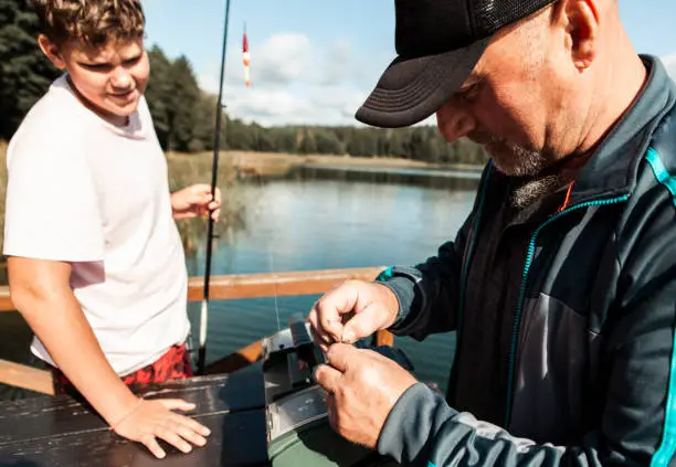 September 19,  2020 - Druskininkai, Lithuania: togetherness and bonding of two generation family men - grandpa is showing and teaching his teenage grandson putting bait on a fishing hook. Both smiling, being happy, cheerful on a sunny day.