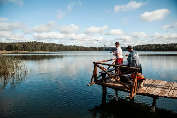 September 19,  2020 - Druskininkai, Lithuania: Grandfather and grandson fishing. Active senior strong man is helping and teaching his teen grandson how to fish. They are standing on a wooden jetty, sunlit, side view.