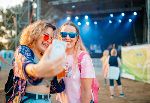 Girlfriends smiling and taking a selfie with mobile phone on daytime outdoor music festival, stage in the background blurred for generic look