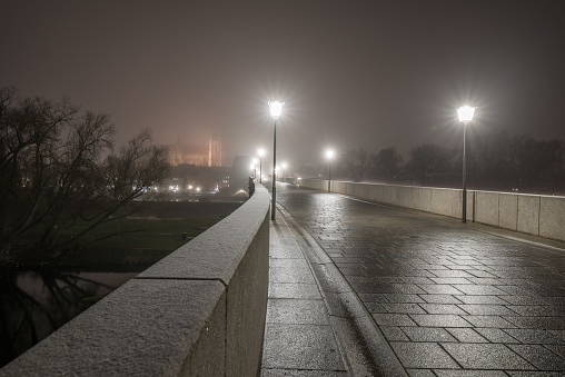 View over snowy stone bridge in Regensburg at night in fog over river Danube with illuminated cathedral in background, Germany 2020