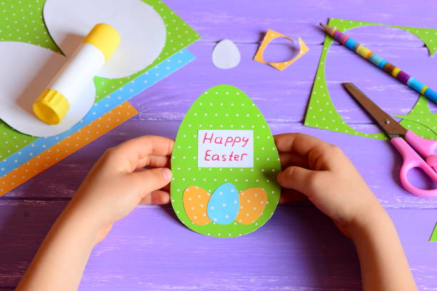 istock Small child holds a Easter greeting card in hands. Child made a Happy Easter greeting card diy in egg shape. Imagination and creativity for kids development. Easter crafts concept. Stationery on a wooden table 1300480475