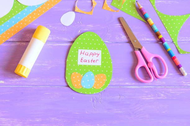 istock Easter greeting card with egg made of colored cardboard, colored sheets, scissors, paper templates in shape of eggs, glue stick, pencil on a wood table. Easy children paper crafts for Easter. Easter handcraft 1300480485