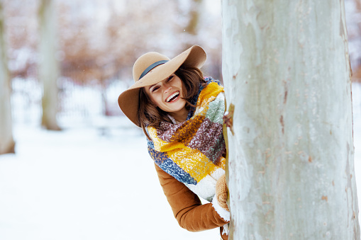 smiling stylish woman in brown hat and scarf with mittens in sheepskin coat near tree outdoors in the city park in winter.