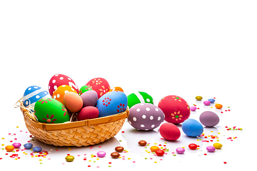 Colorful hand painted Easter eggs arranged in a wicker basket at the bottom of a white background leaving useful copy space for text and/or logo. Sugar sprinkles and candies complete the composition. High resolution 42Mp studio digital capture taken with Sony A7rII and Sony FE 90mm f2.8 macro G OSS lens