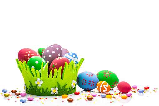 Colorful hand painted Easter eggs arranged in a green basket at the bottom of a white background leaving useful copy space for text and/or logo. Sugar sprinkles and candies complete the composition. High resolution 42Mp studio digital capture taken with Sony A7rII and Sony FE 90mm f2.8 macro G OSS lens
