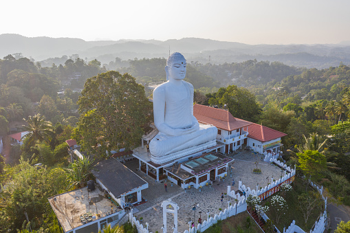 Big white Buddha sitting on top of temple overlooking the green hills around, sunset time. Kandy, Sri Lanka