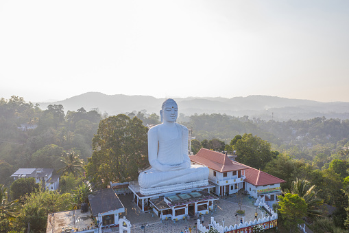 Big white Buddha sitting on top of temple overlooking the green hills around, sunset time. Kandy, Sri Lanka
