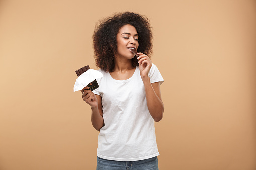 Portrait of a smiling young african woman eating chocolate bar isolated over beige background