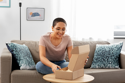 delivery, shipping and people concept - happy young african american woman opening parcel box at home